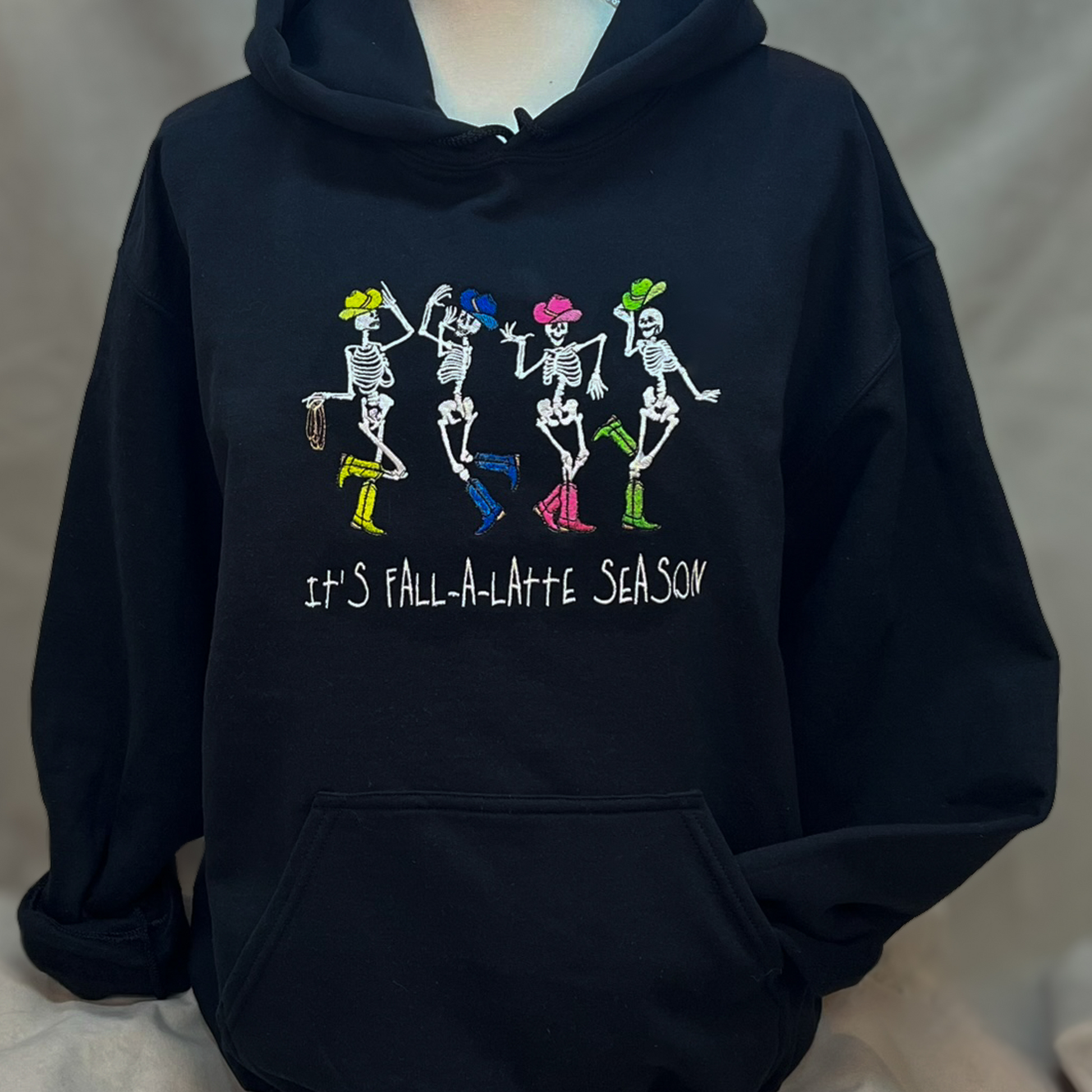 Front view of black glow-in-the-dark skeleton hoodie with “It’s Fall-A-Latte Season” text and dancing cowgirl skeletons